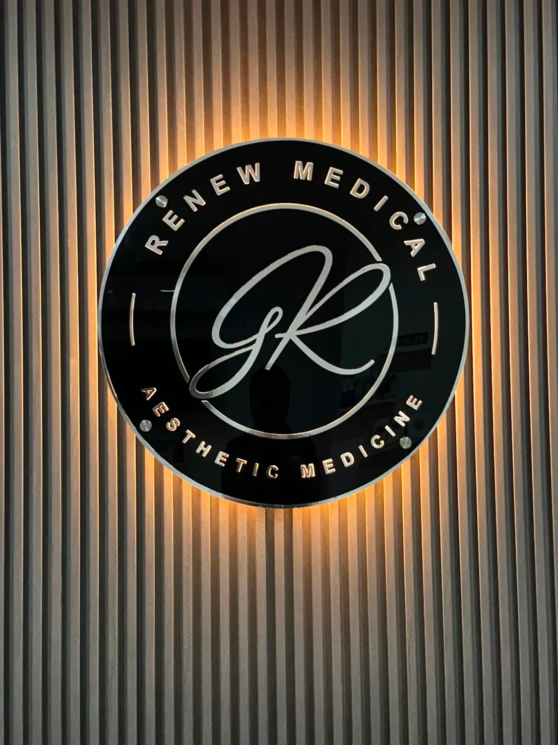 Logotipo circular de Renew Medical en acrílico negro con letras plateadas y luz cálida detrás, sobre un muro de listones